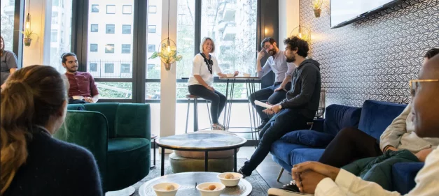 Group of people sitting inside a lounge with large windows, looking out at city buildings. 