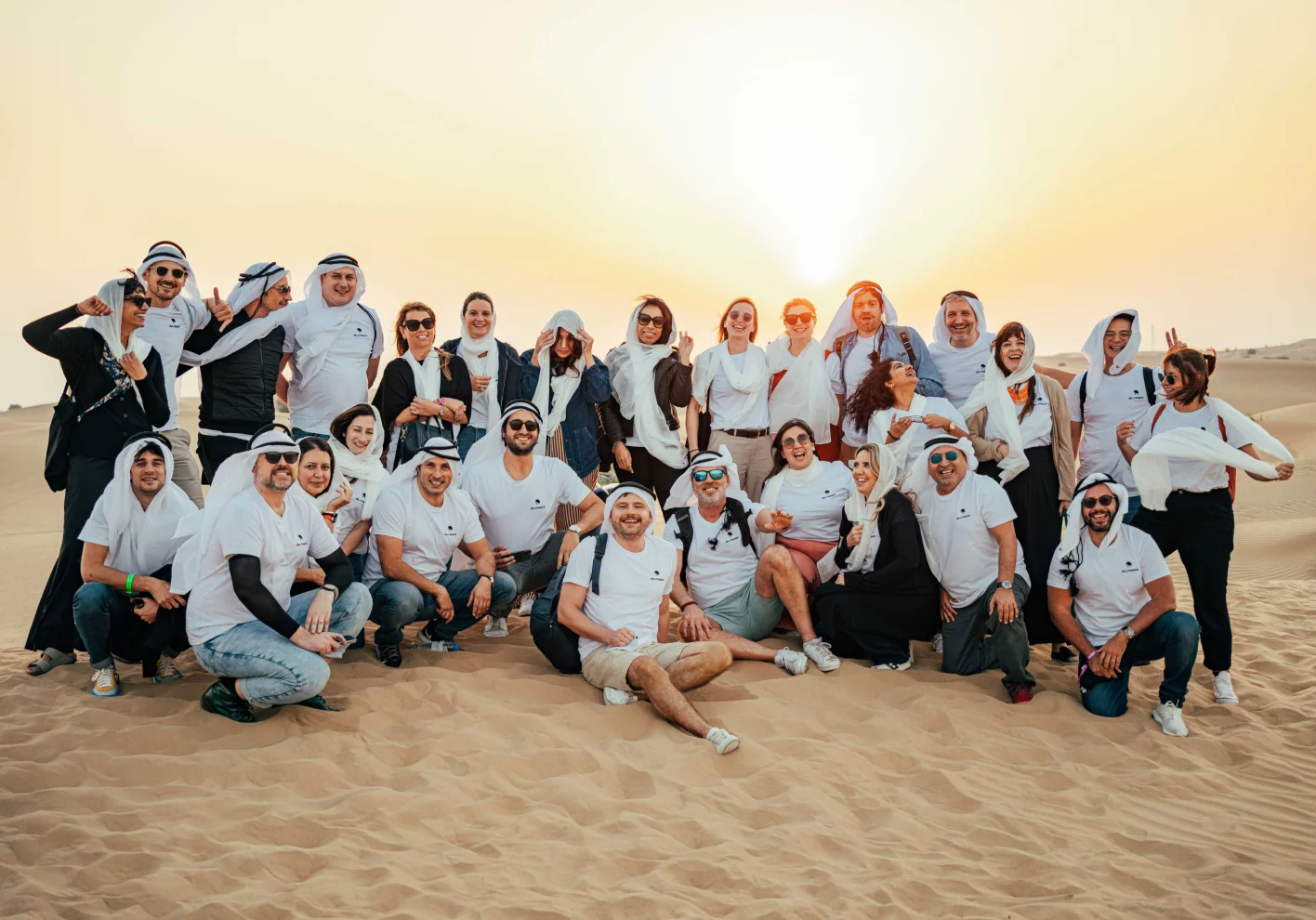 Large group of Jellysmack employees posing in the sand dunes at sunset dressed mostly in white. 