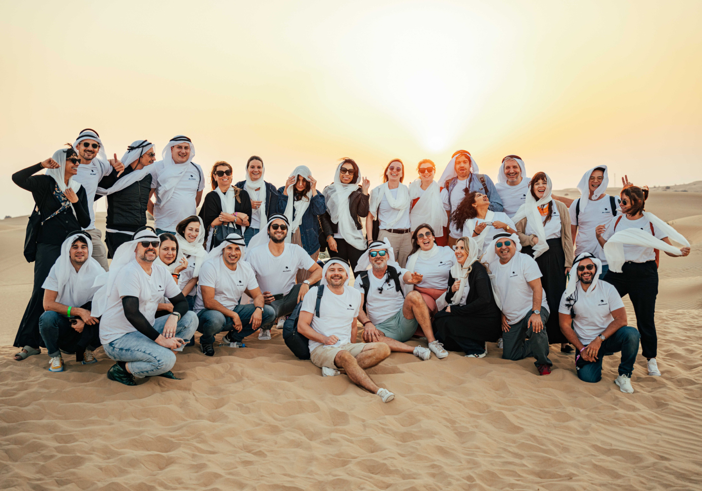 Large group of Jellysmack employees posing in the sand dunes at sunset dressed mostly in white.