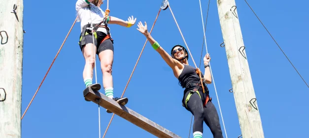 Two female Jellysmack employees high fiving while standing on an elevated wood plank at outdoor ropes course. 
