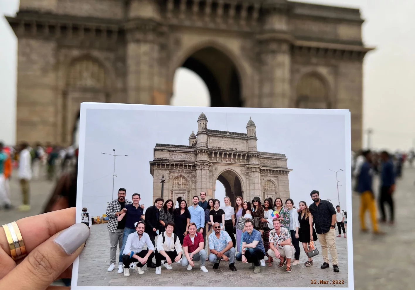 Woman's hand holding a photo of a group of people posing in front of an archway landmark with actual landmark in background.